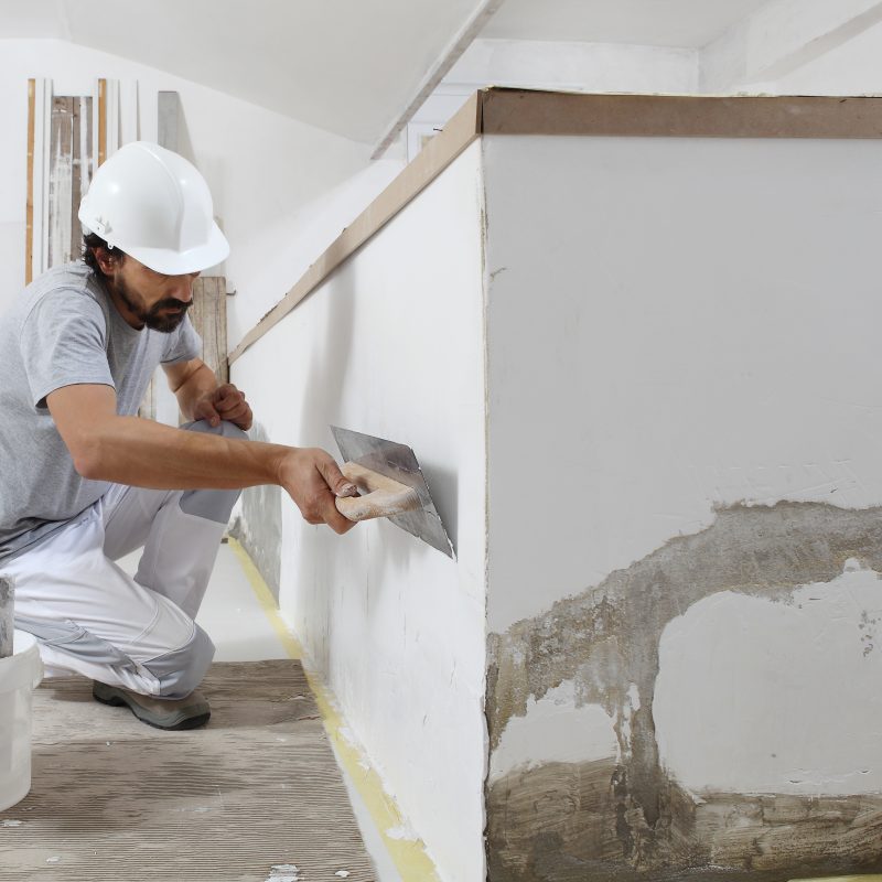 man plasterer construction worker at work, takes plaster from bucket and puts it on trowel to plastering the wall, wears helmet inside the building site of a house